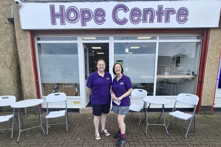 Two members of staff from the Hope Centre community food pantry dressed in purple polo shirts standing outside the front of the Hope Centre shop